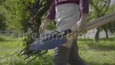 不认识的农民戴着花园手套，手里拿着铲子和草叉，在花园里走来走去。 概念